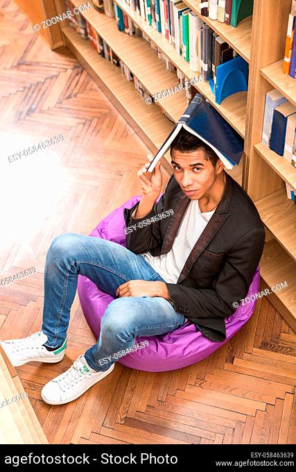 Top view of handsome man keeping book on his head while sitting on the floor and studying in library