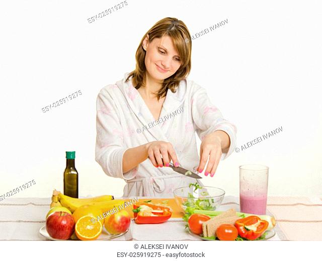 Young beautiful girl in a bathrobe prepares vegetarian salad from vegetables and greens isolated on white background