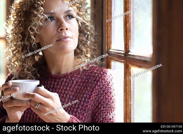Serene people concept lifestyle. One woman drinking coffee or healthy beverage tea alone ner the window at home looking outside and thinking