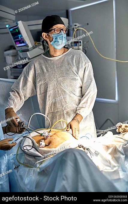 Doctor is using a laparoscopy camera inside the patient's abdominal during the operation. There is an EKG monitor behind them. Vertical