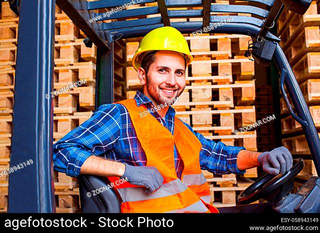 Handsome smiling worker driving forklift in warehouse. Woodworking industry concept. Worker wears protective helmet, vest and gloves