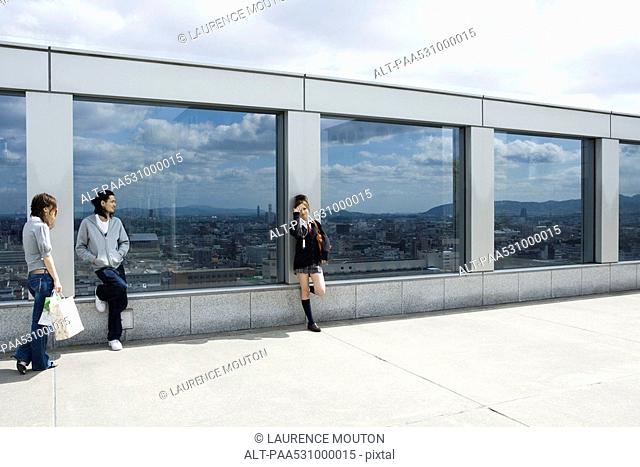 Three young friends leaning against glass wall overlooking city, Kyoto, Japan