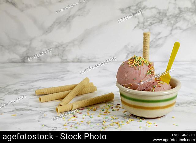 Two strawberry ice cream balls on ceramic bowl with yellow plastic teaspoon and biscuit and various biscuits on marble background. Copy space