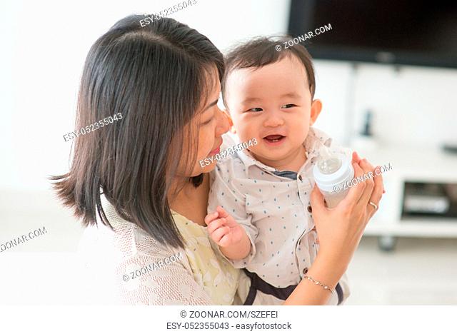 Mother holding milk bottle with happy 9 months old baby. Asian family at home, living lifestyle indoors
