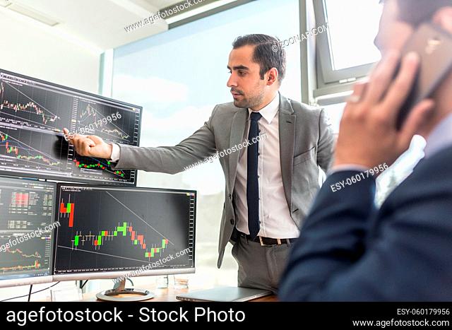Businessmen trading stocks online. Stock brokers looking at graphs, indexes and numbers on multiple computer screens. Colleagues in discussion in traders office