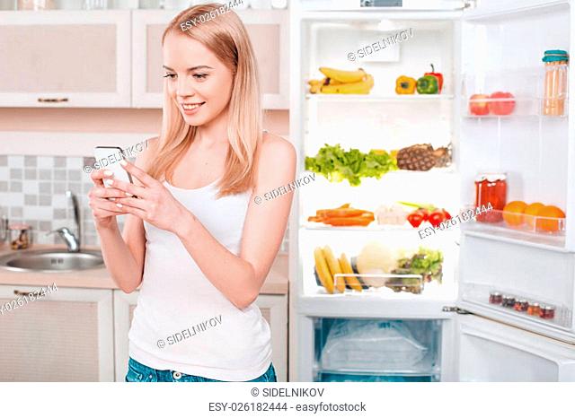Pretty blonde standing near open fridge full of food. Young woman smiling and using mobile phone