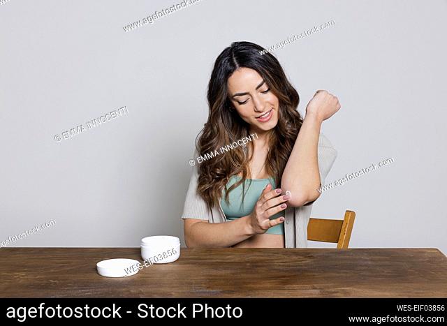 Beautiful woman applying cream on elbow sitting at table against gray background
