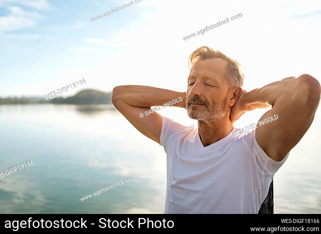 Mature man with eyes closed and hands behind head by lake on sunny day