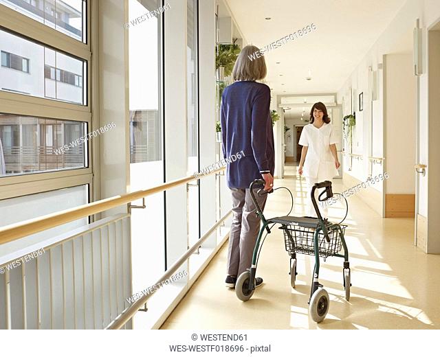 Germany, Cologne, Senior women holding walking frame in corridor, caretaker in background