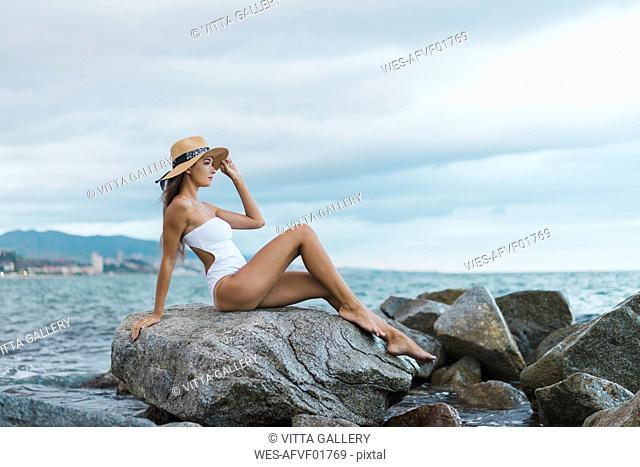 Beautiful young woman wearing swimsuit and hat sitting on rock in the sea