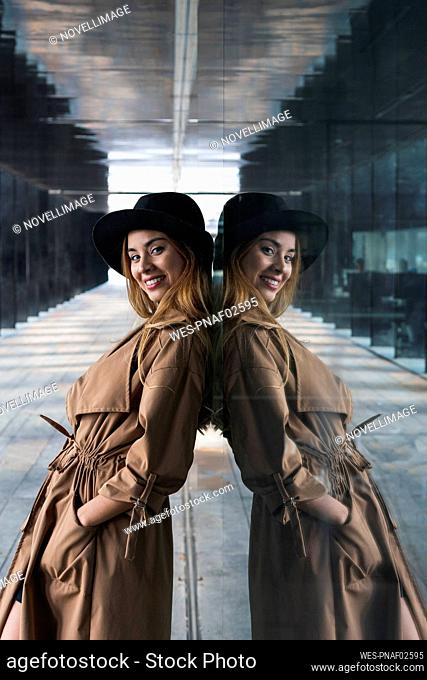 Smiling woman with hand in pocket leaning on glass wall