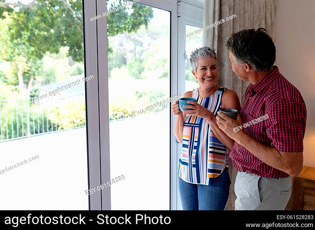 Happy senior caucasian couple standing by window talking holding cups of coffee