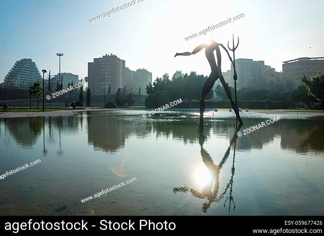 Modern statue of waterman and lightened head with trident in pond at Turia River Park with cityscape, Valencia, Spain