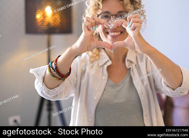 Happy beautiful adult portrait of woman doing heart shape with hands in front of the camera with house indoor background - cheerful female people love and enjoy...