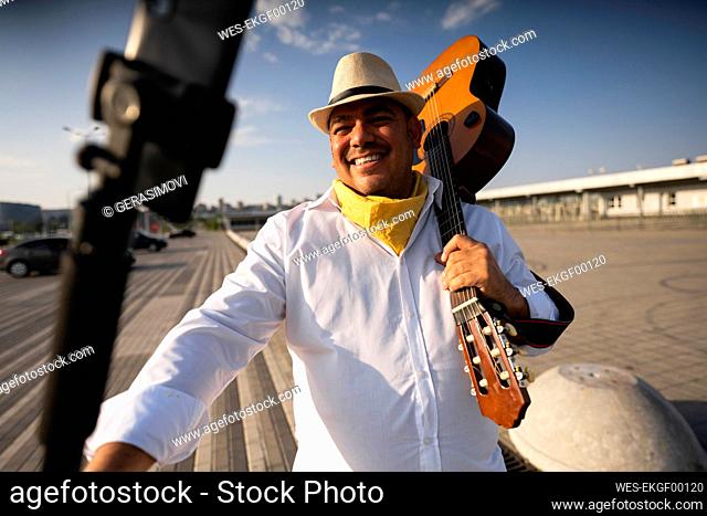 Smiling guitarist holding guitar taking selfie through smart phone on sunny day