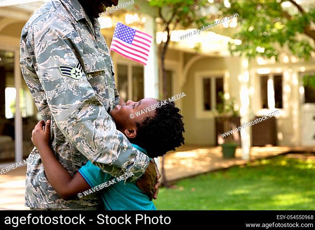 Side view close up of a young adult African American male soldier in the garden outside his home, embracing his young son