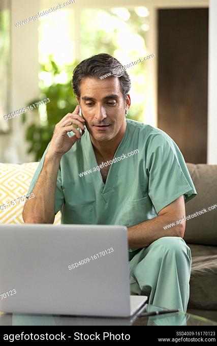 Hispanic Male doctor practicing tele-medicine from his home, using cell phone and laptop computer, Talking to patient on video call