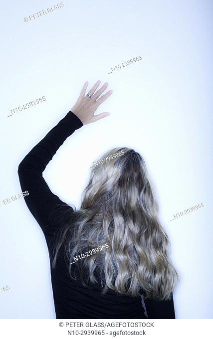 Young blonde woman, with her arm raised, facing a wall