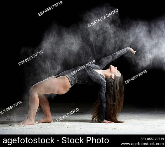 young beautiful caucasian woman with long hair is dressed in a sports black bodysuit stands in a bent pose and throws white flour, black background