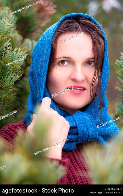 Portrait of brunette woman with long hair and brown eyes standing in pine forest. Pretty young woman dressed in brown knitted pullover and blue scarf thrown...