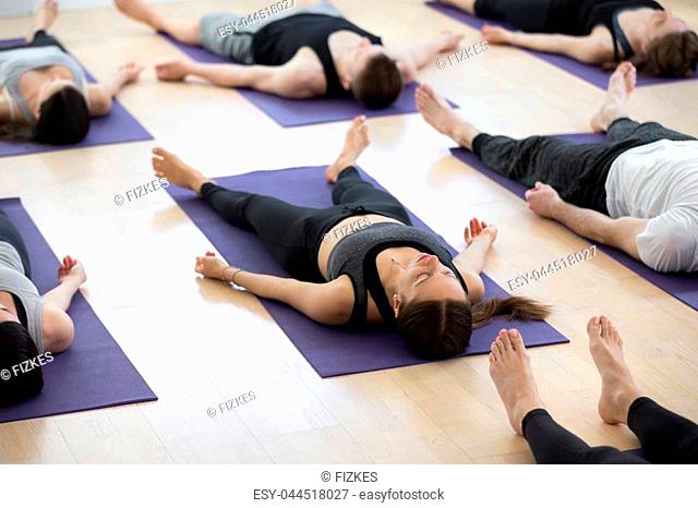 Group of young sporty people practicing yoga lesson, doing Savasana pose, Corpse exercise, Dead Body, working out, indoor full length