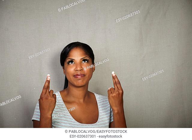 African woman in her 20s praying while crossing her fingers against grey background