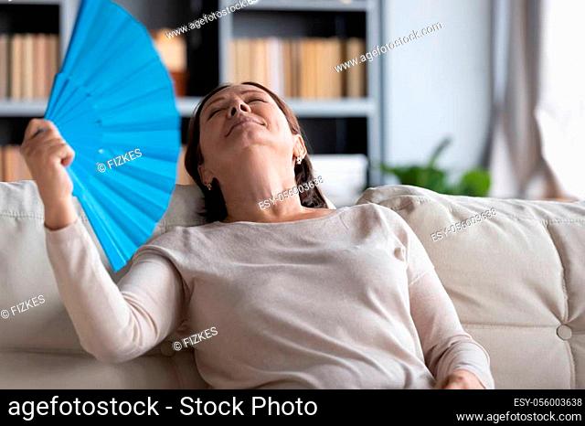Relaxed middle-aged woman sit rest on comfortable couch in living room feel overheated wave with hand fan, senior female relax on cozy sofa feel hot breathe...