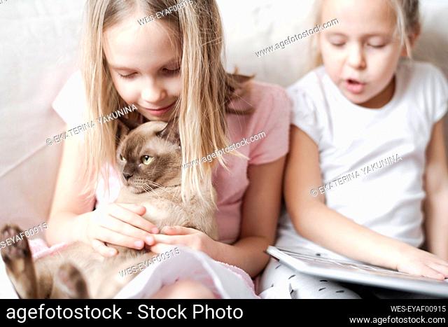Smiling little girl stroking Burmese cat while her sister reading a book