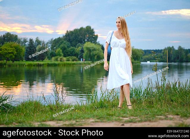 Young woman in long white sundress standing on the river bank during sunset