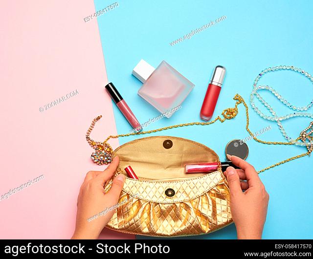 female hands hold a golden clutch bag with various cosmetics and jewelry on a blue background, top view