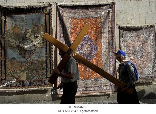 Couple of pilgrims carrying a cross on the Via Dolorosa during Good Friday Catholic procession, walking in front of carpets displayed for sale, Old City