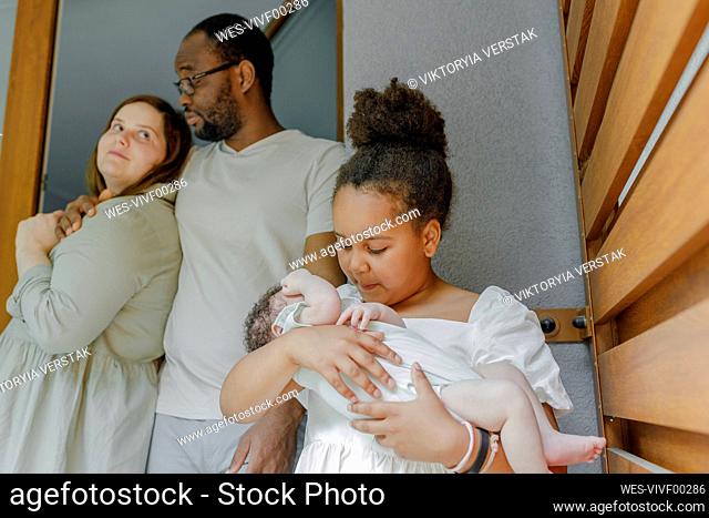 Girl carrying baby girl with parents standing at doorway