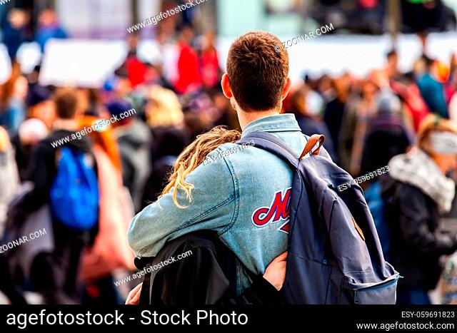 A young millennial couple are seen cuddling from behind, as eco-activists march for the environment in a blurry background, with room for copy