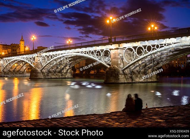 Young couple drinking sparkling wine by the Guadalquivir river and the Triana bridge, Seville, Spain