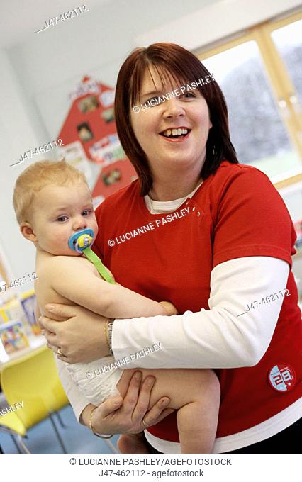 Health visitor holding baby boy in her arms, with dummy in his mouth and wearing only a nappy