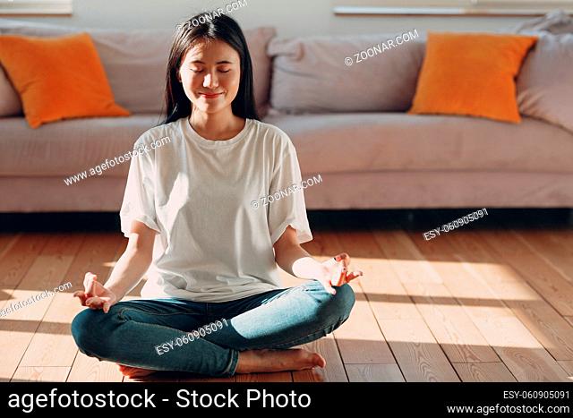 Asian woman doing yoga and zen like meditation lotus pose in casual wear at indoor living room apartment with natural sun light illuminated