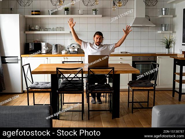 Excited businessman feeling happy about successful project. Man holding hands in the air and looking at laptop while sitting on a kitchen at home
