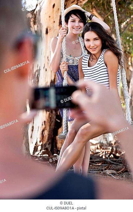 Man taking photograph of two young women sitting on rope swing