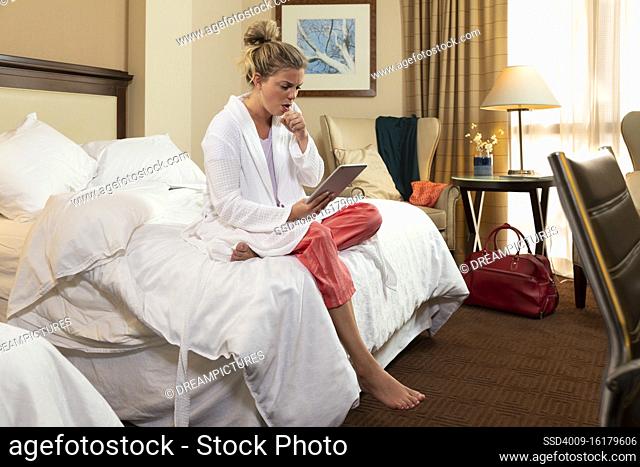 Coughing young Caucasian woman wearing bathrobe in a hotel room, sitting on edge of bed using tablet device to connect with doctor