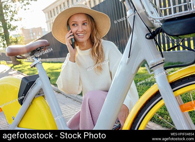 Smiling blond woman talking on smart phone while crouching by bicycle