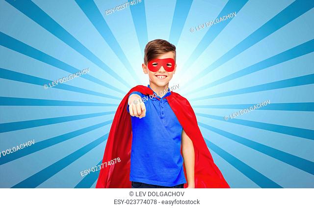 childhood, gesture, carnival costume and people concept - happy boy in red superhero cape and mask pointing finger to you over blue burst rays background