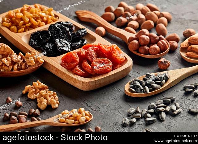 Various varieties of nuts, seeds and dried fruits lying in wooden spoones and dish on black slate background. Healthy food. Vegetarian nutrition