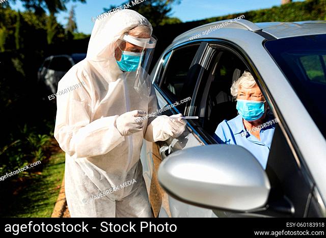 Medical worker wearing ppe suit taking swab test of senior caucasian woman sitting in car