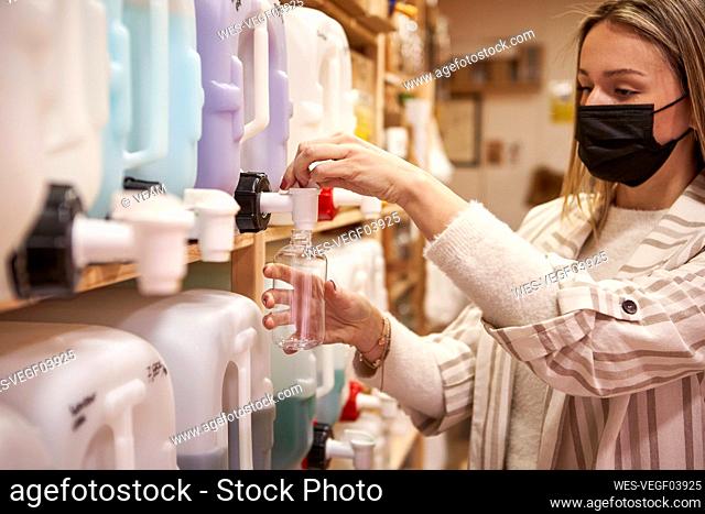 Woman in protective face mask filling bottle from cane at store