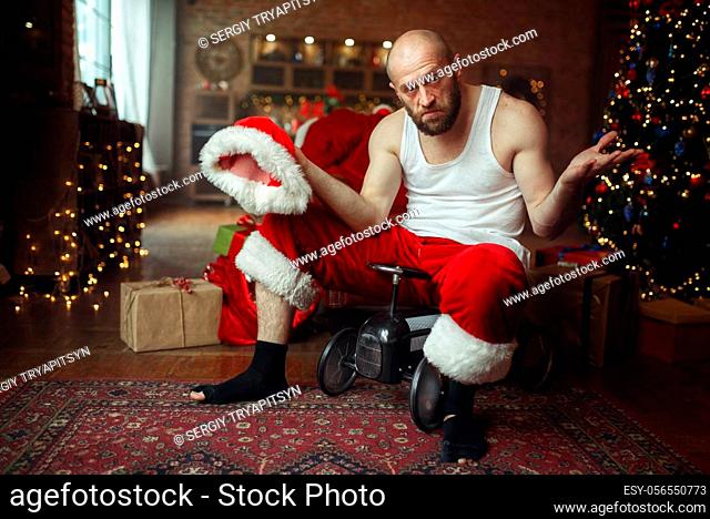 Bad drunk Santa claus in red hat riding on little toy car, nasty party, humor. Unhealthy lifestyle, bearded man in holiday costume, new year