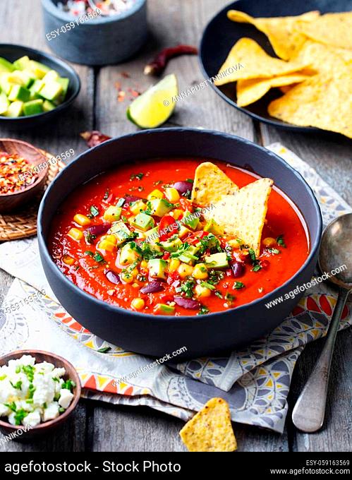 Mexican tomato, bean, bell pepper soup in black bowl with nachos chips. Wooden background