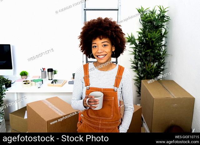 Happy Afro young woman holding coffee cup while standing in new loft apartment
