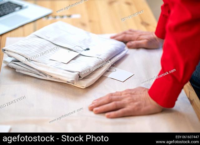 Exclusive shirt. Light-colored shirt with price tag lying folded on white wrapping paper and hands of seller leaning on table