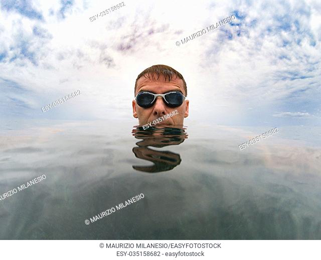 Young serious man in the sea, his head is on the smooth surface of the water, over him the cloudy sky, he wears goggles