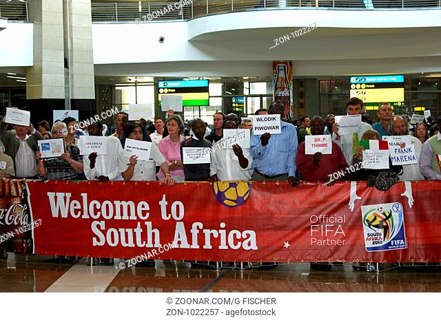 Vertreter von Reiseunternehmen mit Namensschildern in der Hand warten auf ankommende Reisegäste hinter einem Banner mit der Aufschrift Willkommen in Südafrika...